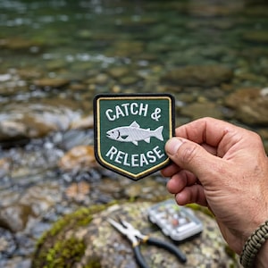 May include: A shield-shaped embroidered patch with a dark green background and a yellow border. The patch features a white fish graphic in the centre with the white text CATCH & above the fish and the white text RELEASE below the fish.