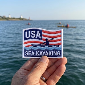 以下が含まれることがあります： 赤と白のストライプと青いフィールドを持つ、様式化されたアメリカ国旗が描かれた長方形の刺繍ワッペン。白い文字でUSAと書かれ、青い波の背景に白い文字でSEA KAYAKINGと書かれており、濃い青色のカヤックのシルエットが中央に配置されています。