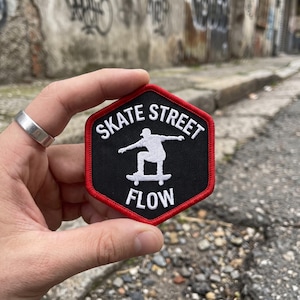 May include: A hexagonal embroidered patch with a black background and a red border. The patch features a white graphic of a person on a skateboard in the center, with the white text SKATE STREET above and the word FLOW below the graphic.