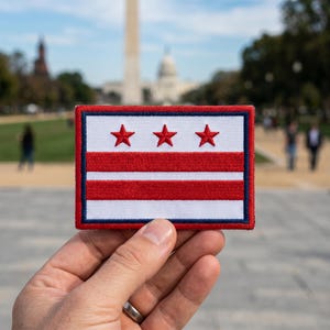 Puede incluir: Un parche rectangular con la bandera de Washington, D.C. El diseño incluye tres estrellas rojas en la parte superior, sobre dos franjas rojas horizontales sobre un fondo blanco, todo enmarcado por un borde azul. El parche está sostenido en una mano.