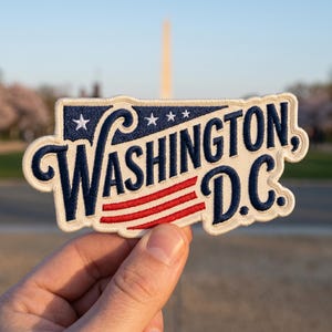 May include: Embroidered patch featuring the text "Washington, D.C." in navy blue script with red stripes and white stars. The patch has a cream-colored border and is held in front of a blurred outdoor background.