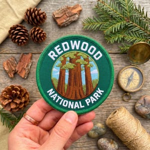 Pode incluir: Um patch verde circular com as palavras "REDWOOD NATIONAL PARK" em letras brancas. O patch apresenta um gráfico de sequoias. O patch é cercado por pinhas, casca e uma bússola.