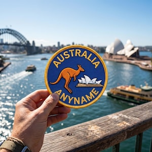 Könnte beinhalten: Ein runder, bestickter Aufnäher mit blauem Hintergrund und gelbem Rand. Der Aufnäher zeigt die Silhouette eines orangefarbenen Kängurus, ein weißes Symbol des Sydney Opera House sowie den Text "AUSTRALIA" oben und "ANYNAME" unten.