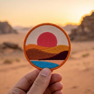 May include: Round embroidered patch featuring a desert landscape with a red sun, orange and brown hills, and a blue body of water. The patch has an orange border and is held in a hand against a desert backdrop.