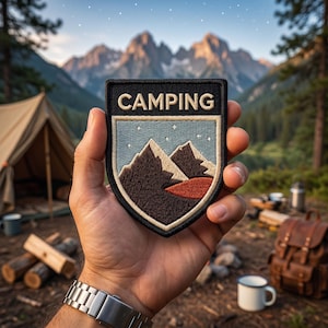 Op de afbeelding: Een zwart en beige embleem met het woord "CAMPING" bovenaan. Het embleem toont een berglandschap met een blauwe lucht en sterren. Het embleem wordt vastgehouden voor een berg- en bosachtergrond.