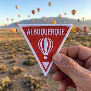 May include: A triangular embroidered patch with a red background and white border. The patch features the white text ALBUQUERQUE above a white graphic of a hot air balloon and a small white triangle at the bottom point of the patch.