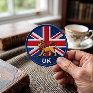 Op de afbeelding: Een ronde geborduurde patch met de Union Jack vlag in rood, wit en blauw. Een goudbruine leeuw staat in het midden en de letters UK zijn onderaan in het wit geborduurd. De patch heeft een donkerblauwe rand en strakke stiksels.