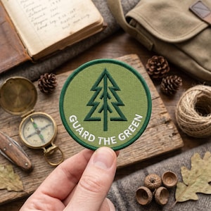 Könnte beinhalten: Ein runder, grüner Aufnäher mit einem dunkelgrünen Kieferndesign und den Worten "GUARD THE GREEN" in Weiß. Der Aufnäher wird in einer Hand gehalten, mit einem Kompass, einem Tagebuch und anderen Outdoor-Artikeln im Hintergrund.