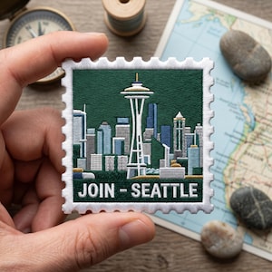 May include: Embroidered patch featuring the Seattle skyline and the Space Needle, with the text "JOIN - SEATTLE" in white. The patch is green with a white border, held in a hand. Other items in the background include a compass and a map.