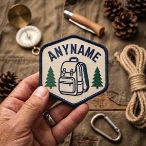 Może przedstawiać: Beżowa i granatowa naszywka z napisem "ANYNAME" nad ilustracją plecaka i dwoma zielonymi sosnami. Naszywka jest trzymana w dłoni, z kompasem, nożem, liną i szyszkami w tle.