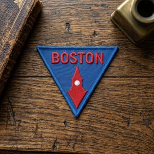 Pode incluir: Um emblema triangular bordado com fundo azul e a palavra BOSTON costurada em letras vermelhas. Abaixo do texto, há um ícone de torre vermelha estilizada com um pequeno detalhe circular branco no centro, contornado por costura azul.
