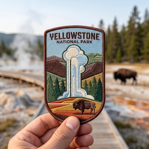 Op de afbeelding: Geborduurde patch met de tekst "YELLOWSTONE NATIONAL PARK". De patch toont een geiser, een bizon, bergen en bomen in blauw, groen, bruin en oranje. De patch wordt in de hand van een persoon gehouden.