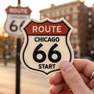 May include: A Route 66 Chicago patch with a cream background, black text, and a red border. The patch reads "ROUTE CHICAGO 66 START". A Route 66 sign is in the background.
