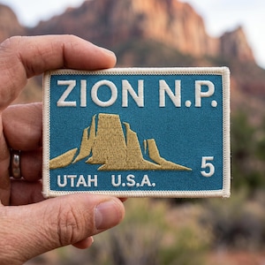 Peut inclure: Écusson rectangulaire bleu avec le texte blanc "ZION N.P." au-dessus d'une illustration de montagne dorée. Le texte "UTAH U.S.A." et le chiffre "5" sont en dessous. Bordure blanche.