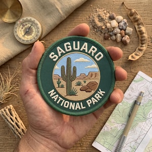 May include: A round, green embroidered patch with the text "SAGUARO NATIONAL PARK" and a desert landscape design. The patch features a large cactus, mountains, and blue sky. The patch is held in a hand.