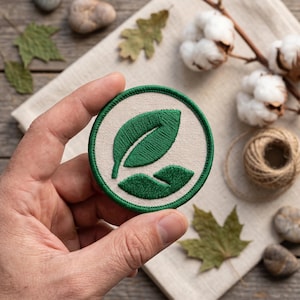 Può includere: Una toppa rotonda ricamata con un disegno a foglia verde su sfondo color crema. La toppa è tenuta in mano, con un bordo verde. L'immagine include batuffoli di cotone, spago e foglie secche.