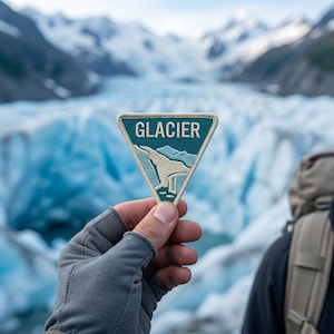 May include: A triangular embroidered patch featuring a teal background with the word GLACIER in cream-colored lettering above a stylized cream-colored mountain and glacier graphic, all bordered by a cream-colored stitched edge.