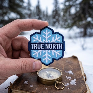 May include: A blue and white embroidered patch with the text "TRUE NORTH" and a snowflake design. A vintage brass compass rests on a weathered leather-bound book. The background features a snowy forest scene.