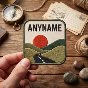 Op de afbeelding: Geborduurde patch met de tekst "ANYNAME" boven een landschap. De scène toont een rode zon, groene heuvels en een kronkelende weg. De patch heeft een crèmekleurige achtergrond en een groene rand.