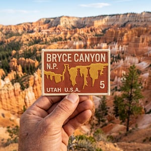 Peut inclure: Écusson rectangulaire avec le texte "BRYCE CANYON N.P. UTAH U.S.A." en lettres blanches et dorées. L'écusson présente une illustration dorée du canyon et le chiffre 5. L'écusson est tenu devant une vue panoramique de Bryce Canyon.