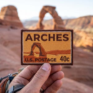 Peut inclure: Écusson brodé avec le mot "ARCHES" et une illustration d'une arche en grès, rappelant un timbre-poste. L'écusson a une bordure marron et le texte "U.S. POSTAGE 40c". L'arrière-plan montre une vue floue d'une arche naturelle.