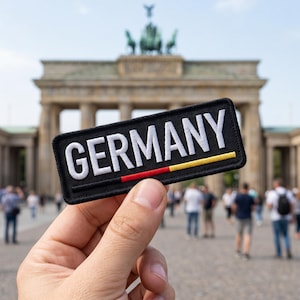 Puede incluir: Un parche rectangular negro con la palabra "GERMANY" en letras blancas. El parche presenta un diseño de rayas rojas, amarillas y negras, sostenido frente a la Puerta de Brandeburgo en Berlín.