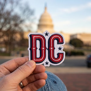 May include: Embroidered patch featuring the letters "DC" in red, white, and blue, with star accents. The patch is held in front of the U.S. Capitol Building, suggesting a Washington D.C. souvenir or travel item.