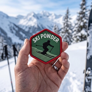 May include: A hexagonal embroidered patch with a red border, a dark green background, and a light green lower section. The patch features a black silhouette of a skier and the white text "SKI POWDER" arched across the top portion.