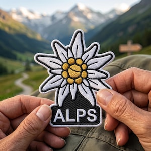 May include: A white and yellow edelweiss flower patch with black trim and the word "ALPS" in white letters on a black background. The patch is held up against a backdrop of mountains and a green valley.