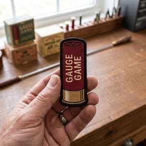 May include: A rectangular embroidered patch featuring a maroon and tan shotgun shell design with a black border. The text GAUGE GAME is stitched in tan lettering vertically along the center of the maroon section of the shell graphic.