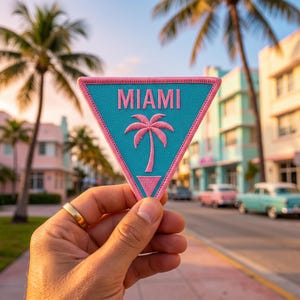 May include: A triangular embroidered patch featuring a bright turquoise background with a pink border. The word MIAMI is stitched in pink capital letters above a pink palm tree icon, with a small pink triangle shape at the bottom point.