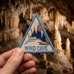 May include: A triangular embroidered patch with a light blue border. The design features a beige and dark blue cave interior with stalactites and stalagmites. The text WIND CAVE is embroidered in blue capital letters at the bottom of the patch.