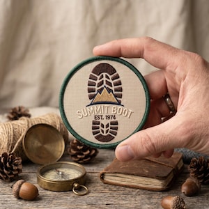 May include: Round patch with a green border and the words "SUMMIT BOOT EST. 1974". The patch features a brown boot print with a mountain graphic in the center. The patch is held by a hand, with a compass, journal, and pinecones in the background.