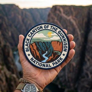 Può includere: Una toppa rotonda con la scritta "Black Canyon of the Gunnison National Park". La toppa raffigura un paesaggio del canyon con un fiume, il sole e le nuvole. La toppa è tenuta in mano.