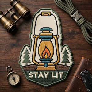 May include: Embroidered patch featuring a vintage lantern with a flame, two green pine trees, and the text "STAY LIT." The patch has a cream-colored background and a green border. Other items include a compass, binoculars, and a leather-bound notebook.