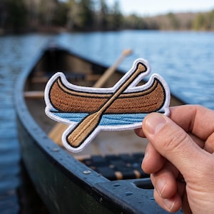 Könnte beinhalten: Gestickter Aufnäher mit einem braunen Kanu- und Paddeldesign mit weißem Rand. Der Aufnäher wird vor einem Kanu auf einem See gehalten. Der Kanu-Aufnäher ist ein tolles Accessoire für Outdoor-Enthusiasten.