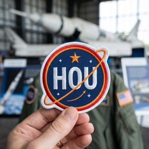 Könnte beinhalten: Gestickter Aufnäher mit dem Wort "HOU" in Weiß, umgeben von einem blauen Kreis mit orangefarbenem Rand und einem goldenen Stern. Der Aufnäher hat ein planetenähnliches Design mit einer orangefarbenen Orbitallinie. Eine Hand hält den Aufnäher.