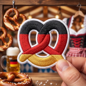 Può includere: Toppa ricamata a forma di pretzel, con i colori della bandiera tedesca: nero, rosso e oro. La toppa è mostrata su uno sfondo di legno con pretzel, birra e abbigliamento tradizionale tedesco.