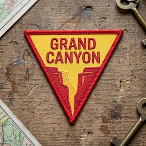 Puede incluir: Un parche triangular bordado con fondo amarillo y el texto rojo GRAND CANYON. El diseño incluye un gráfico estilizado de un cañón rojo que divide el campo amarillo, todo bordeado por un grueso borde cosido en rojo.