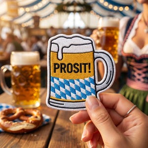 Op de afbeelding: Geborduurde patch in de vorm van een bierpul, met het woord "PROSIT!" in het zwart. De pul heeft een geel bovenstuk, een blauw en wit ruitpatroon en een witte schuimlaag. Een hand houdt de patch voor een vage achtergrond.