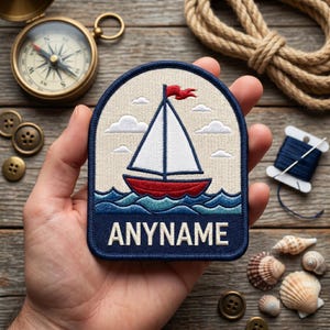Op de afbeelding: Geborduurde patch met een zeilboot met een wit zeil, rode romp en blauwe golven. De patch heeft een marineblauwe rand en de tekst "ANYNAME" in het wit. Een rode vlag wappert van de mast. De patch wordt in een hand gehouden.