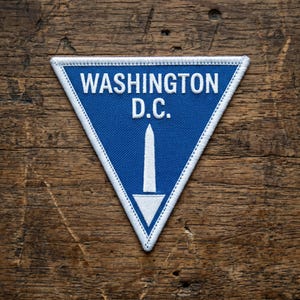 Puede incluir: Un parche triangular bordado con fondo azul y borde blanco. El texto WASHINGTON D.C. está bordado en letras mayúsculas blancas sobre una representación gráfica blanca del Monumento a Washington que apunta hacia abajo.