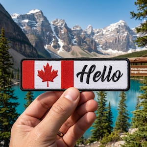 Peut inclure: Écusson rectangulaire avec le drapeau canadien, une feuille d'érable rouge et le mot « Hello » en noir. L'écusson est présenté devant un paysage de montagnes et de lac.