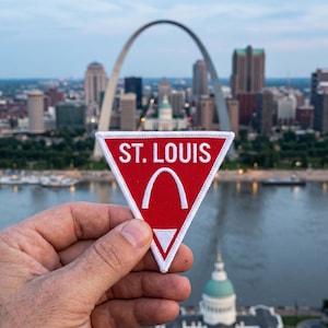 Pode incluir: Um emblema triangular bordado com fundo vermelho e borda branca. O texto ST. LOUIS está bordado em branco na parte superior, com um gráfico branco do Gateway Arch posicionado abaixo do texto e um pequeno triângulo branco na base.
