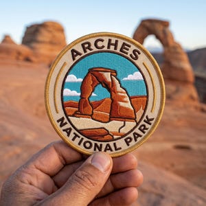 Pode incluir: Patch bordado com o Delicate Arch do Parque Nacional Arches. O patch tem uma borda bege com as palavras "ARCHES NATIONAL PARK" em um design circular. A imagem interna mostra o arco em tons de laranja e marrom contra um céu azul.