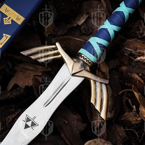 Pode incluir: Uma espada de fantasia com uma l&acirc;mina prateada e detalhes dourados. O punho &eacute; envolto em couro azul e verde-azulado. A bainha &eacute; azul com elementos decorativos dourados. A espada apresenta um s&iacute;mbolo triangular preto e branco na l&acirc;mina.