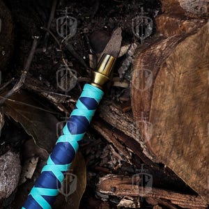 Pode incluir: Um close-up de uma faca decorativa com cabo de lat&atilde;o e punho enrolado em azul e verde-azulado. A faca &eacute; colocada contra um fundo de madeira e folhas, criando uma est&eacute;tica r&uacute;stica.