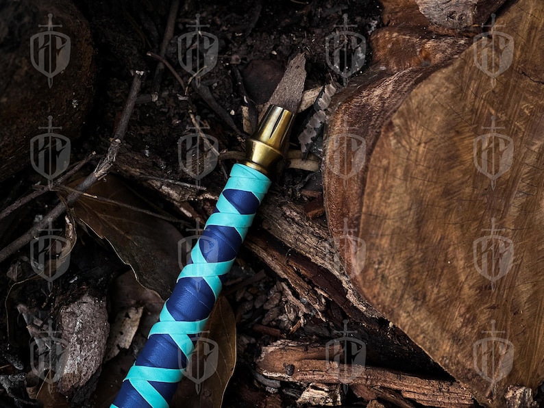 Pode incluir: Um close-up de uma faca decorativa com cabo de lat&atilde;o e punho enrolado em azul e verde-azulado. A faca &eacute; colocada contra um fundo de madeira e folhas, criando uma est&eacute;tica r&uacute;stica.