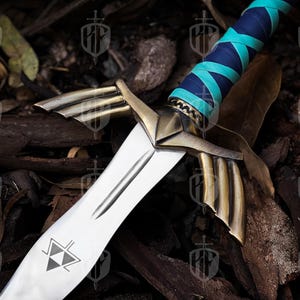 Pode incluir: Uma espada decorativa com uma l&acirc;mina prateada e detalhes dourados. O punho &eacute; envolto em couro azul e verde-azulado. A l&acirc;mina apresenta um design triangular. A espada est&aacute; sobre um fundo de madeira e folhas.