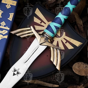 Pode incluir: Uma espada decorativa com uma l&acirc;mina branca e detalhes dourados, com uma empunhadura enrolada em azul e verde-azulado. Uma bainha azul combinando com desenhos dourados e um escudo preto com um emblema dourado tamb&eacute;m s&atilde;o vis&iacute;veis.